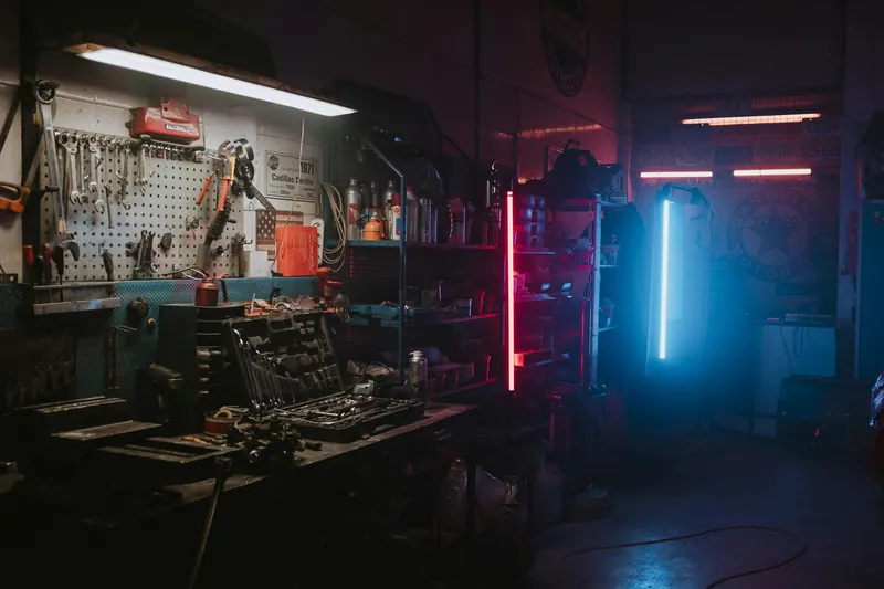 Dim workshop with hand tools on a pegboard wall, shelves of supplies and red and blue neon tube lights